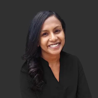 Indu Sudhakar, wearing a black formal top, against a grey backdrop.